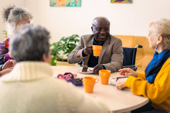 A small group of retired people are sitting around a table in a nursing home, drinking coffee from mugs and eating cake and biscuits