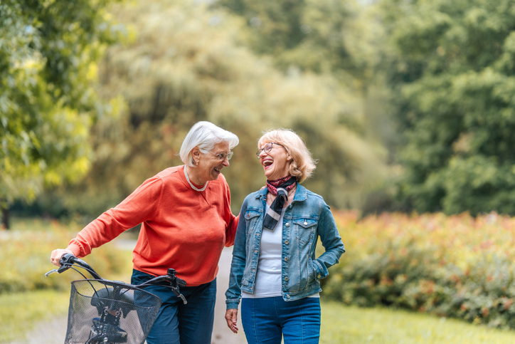 Two joyful elderly Caucasian women enjoy a leisurely walk in a lush park, one friend casually pushing a bicycle, both dressed in casual outdoor wear.